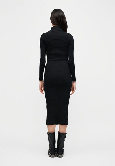 Μαύρο midi φόρεμα ζιβάγκο με μακριά μανίκια, εφαρμοστή σιλουέτα και ριγωτή υφή· φορεμένο με μαύρες μπότες μέχρι το γόνατο με ασημένιες λεπτομέρειες.