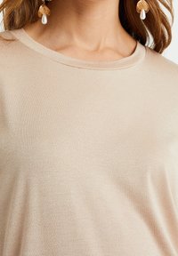 Světle béžový, krátkozákonný top vyrobený z měkké, texturované látky s kulatým výstřihem. Má hladký a mírně lesklý povrch.