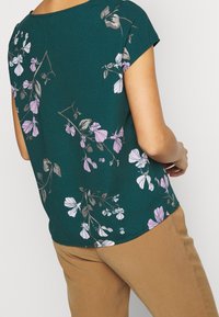 Top verde a maniche corte con motivo floreale in viola e bianco. Tessuto morbido con un fit rilassato e orlo curvo.