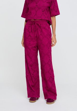 Helle pinke Spitzen-Hose mit weitem Bein, elastischem Bund und Kordelzug, getragen mit passendem Oberteil und pinken Turnschuhen, vor einfachem Hintergrund.