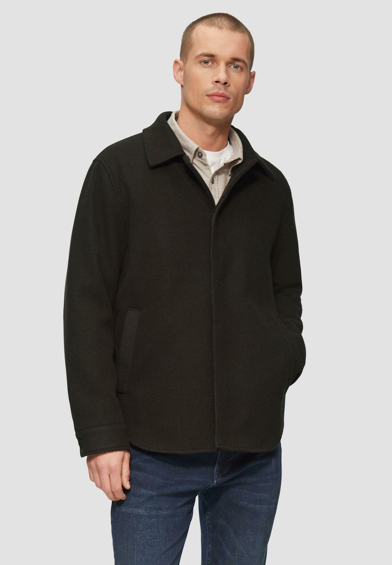 Dunkelgraue Wollmischjacke mit Kragen, Fronttaschen und geradem Schnitt. Getragen über einem Hemd mit Knöpfen, kombiniert mit blauen Jeans.