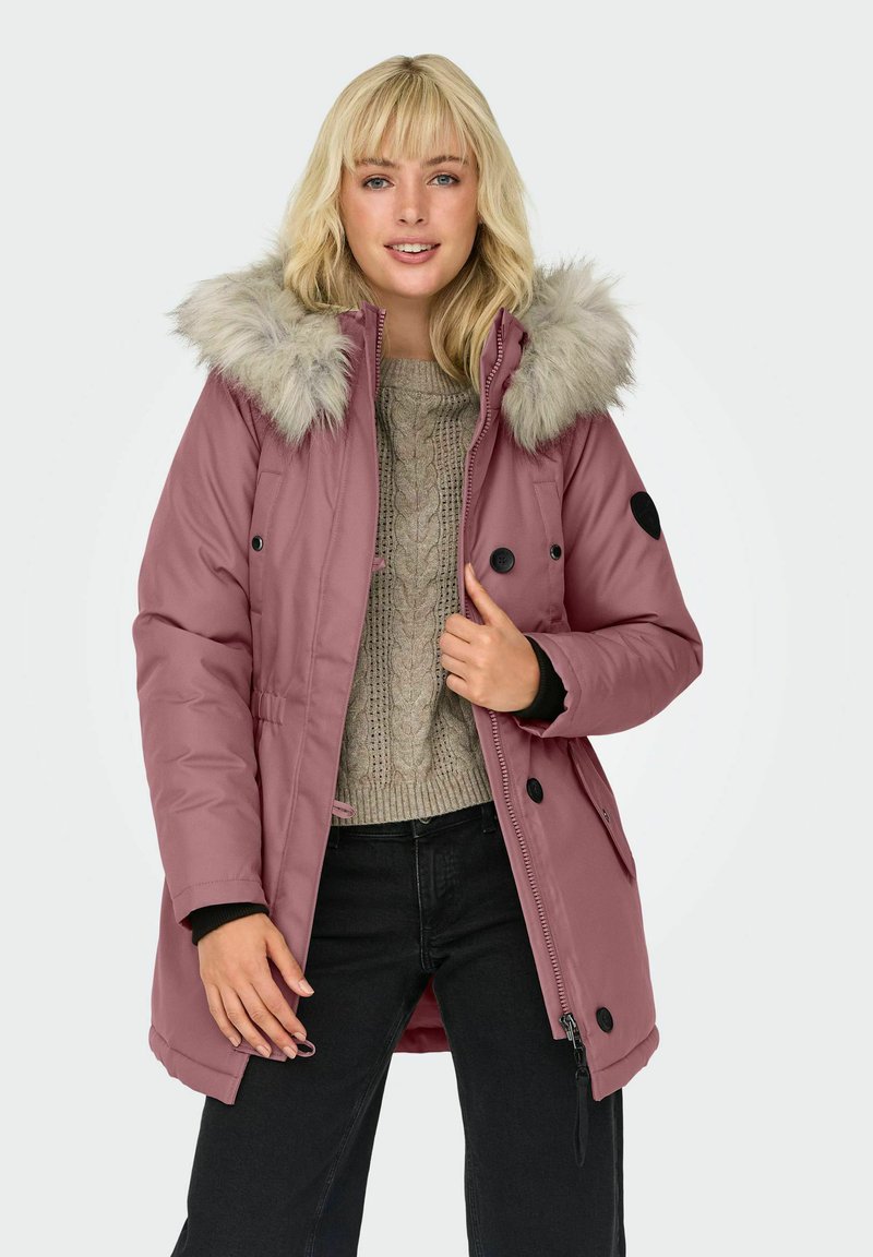 Cappotto imbottito rosa con cappuccio foderato in pelliccia sintetica, caratterizzato da bottoni neri e dettagli neri. Indossato sopra un maglione chiaro in maglia e pantaloni neri.