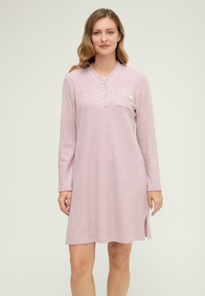 Mujer con cabello ondulado que lleva una camisón de punto waffle de manga larga en rosa claro con botones y un bolsillo en el pecho, de pie contra un fondo liso.