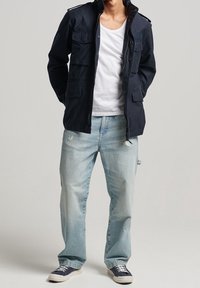 Marineblauwe utility-jas met meerdere zakken, gedragen over een wit T-shirt, gecombineerd met lichtblauwe jeans en marineblauwe sneakers.