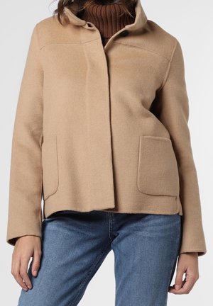 Kvinde iført en beige uldfrakke med store forlommer over en brun rullekrave og blå jeans mod en ensfarvet baggrund.