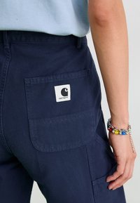 Pantalons cargo bleu marine avec une poche arrière carrée arborant un patch logo Carhartt blanc. Tissu lisse avec des coutures renforcées.