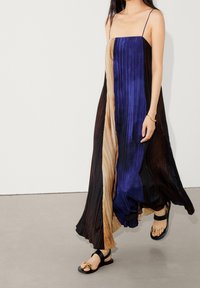 Kleid aus plissiertem Stoff in Blau, Schwarz und Beige; dünne Spaghetti-Träger; knöchellanges Design; getragen mit schwarzen Sandalen.