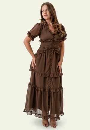 Mujer con cabello largo y marrón que lleva un vestido maxi marrón de capas con mangas abullonadas y botones, de pie con las manos en las caderas.