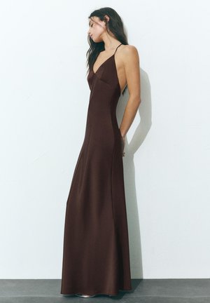 Femme en robe longue marron foncé sans manches avec de fines bretelles, debout de profil contre un mur blanc uni.