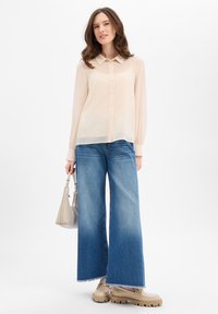 Przezroczysta, jasnobeżowa koszula na guziki z kołnierzykiem, zestawiona z szerokimi, niebieskimi dżinsami o szerokich nogawkach oraz beżowymi butami na masywnej podeszwie. Trzyma małą szarą torebkę.