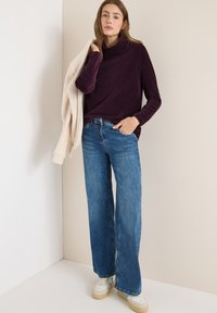 Weite blaue Denim-Jeans kombiniert mit einem tiefpurpurfarbenen, langärmeligen Oberteil mit hohem Kragen. Ein hellbeiger Pullover liegt über einem Arm.