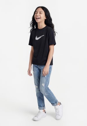 Κορίτσι που γελάει, φοράει μαύρο μπλουζάκι Nike, σκισμένο μπλε τζιν και λευκά αθλητικά παπούτσια, στέκεται μπροστά σε ένα λευκό φόντο.