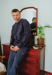 Námořnická modrá mikina s malým oranžovým logem slona, spárovaná s odpovídajícími tepláky s oranžovými akcenty. Umístěno proti dřevěné komodě.