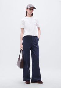 Λευκό βαμβακερό μπλουζάκι με μικρό λογότυπο, σκούρο μπλε παντελόνι με φαρδιά πόδια, γκρι καπέλο, κρατώντας καφέ δερμάτινη τσάντα ώμου και φοράει σκούρα παπούτσια.