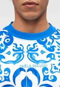 Κοντινή λήψη του κάτω μέρους του προσώπου και του λαιμού ενός ατόμου που φοράει ένα πουκάμισο λευκό και μπλε με μοτίβο, στο οποίο είναι κεντημένο το όνομα «Carlo Colucci» στο στήθος.