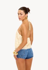 Femme aux cheveux bruns en chignon portant un haut à sequins crème sans manches, un short en denim bleu, des boucles d'oreilles en forme d'étoile dorées et de larges bracelets en or.