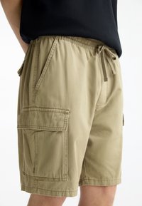 Pantalones cortos de carga beige con cintura elástica, cordón de ajuste y múltiples bolsillos. La tela parece suave con una textura ligera.