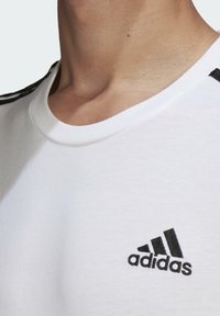 Witte T-shirt met zwart Adidas-logo, met korte mouwen en een ronde hals. Het materiaal lijkt glad en lichtgewicht, met zijstrepen.