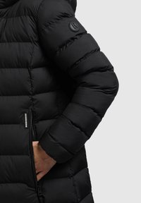 Fekete puffermellény, steppelt mintázattal, cipzáras zsebekkel és kerek logóval a hónalj részen. Puha tapintás és testhezálló forma.