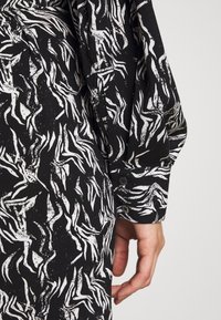 Chemise en tissu noir avec motif abstrait blanc, dotée d'une manche retroussée et de détails à boutons, présentant une apparence fluide et texturée.