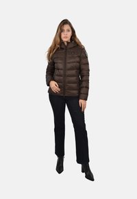 Hnedá prešívaná bunda s kapucňou, vertikálnou prešívanou textúrou, predným zipsom a detailom s nášivkou na ľavom pleci. Vhodná s tmavými džínami a čiernymi čižmami.