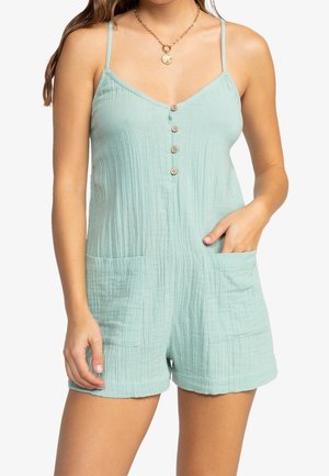 Woman wearing light blue sleeveless romper with buttons down front, two front pockets, and a gold necklace with pendant.