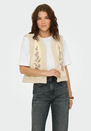 Gilet crema con design floreale ricamato, texture morbida, apertura frontale, indossato sopra una t-shirt bianca e abbinato a jeans scuri.