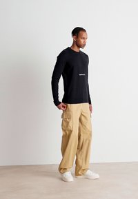 Maglietta nera a maniche lunghe con logo bianco, abbinata a pantaloni cargo beige con tasche laterali e sneaker bianche su uno sfondo neutro.