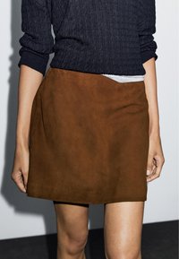 Brauner Minirock aus Wildleder mit glatter Textur, seitlichem Schlitz und sauberem, minimalistischem Design; kombiniert mit einem dunkelblauen Strickpullover.