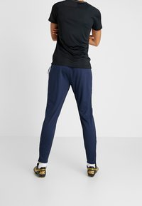 Svart träningsskjorta och marinblå byxor, gjorda av mjukt material. Byxorna har sidofickor och en avsmalnande passform. Skorna är svarta och guldiga fotbollsskor.