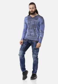 Langärmliges blaues Batikshirt mit V-Ausschnitt und strukturierten Jeans aus Denim, die abwechslungsreiche Patches und Cargo-Taschen aufweisen, kombiniert mit schwarzen Sneakers.