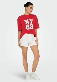 Rode oversized T-shirt met witte "NY 83" opdruk, gecombineerd met witte shorts en witte sneakers, voorzien van ribgebreide manchetten en enkelkousen.
