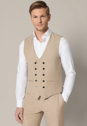 Mężczyzna ubrany w beżową dwurzędową kamizelkę i pasujące spodnie z białą koszulą, stojący z jedną ręką w kieszeni na jednolitym tle.