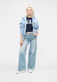 Personne portant un sweat à capuche zippé bleu clair, un t-shirt bleu marine avec le logo GAP, un jean clair à jambes larges et des baskets compensées blanches, debout avec une main dans la poche.