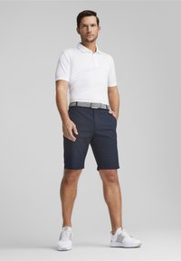 Bela polo majica, mornarsko modre kratke hlače in vzorčast pas. Model nosi bele športne čevlje, stoji s potezanim rokam v žepu. Preprost ozadje.