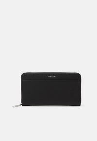 Crna kožna novčanica s teksturiranom površinom, s zatvaranjem na patentni zatvarač i srebrnim Calvin Klein logom sprijeda. Kompaktni i pravokutni dizajn.