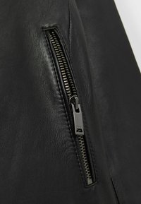 Gros plan d'une veste en cuir noire présentant une fermeture éclair métallique, une surface texturée et un design élégant avec une poche zippée sur le côté.