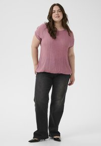 Krøllet woman iført en lyserød kortærmet top med sølvstriber, sorte jeans og sorte spidse hæle, stående mod en hvid baggrund.