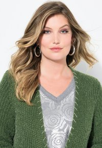 Grön texturerad cardigan med öppen front och vita sömdekorationer, lager på lager över en grå v-ringad topp med ett ros-mönster.