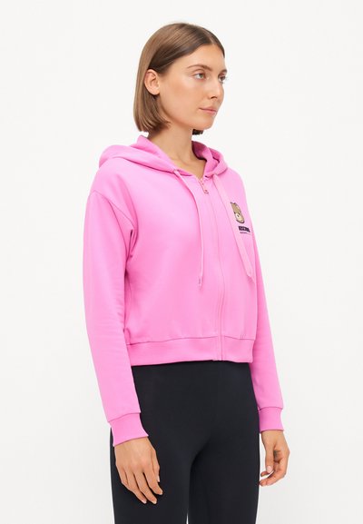 Rožna skrajšana kapuca s poudarkom na zadrgo, izdelana iz mehkih materialov, z grafikami medvedka na levem prsnem delu in vrvicami na kapuci.