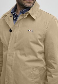 Veste beige en coton avec un col, un bouton noir et un petit logo tricolore sur la poitrine. Texture lisse, design décontracté.