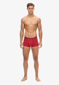 Boxers en coton rouge avec un logo "SUPERDRY" noir sur la ceinture élastique. Présente un ajustement serré et une texture lisse.