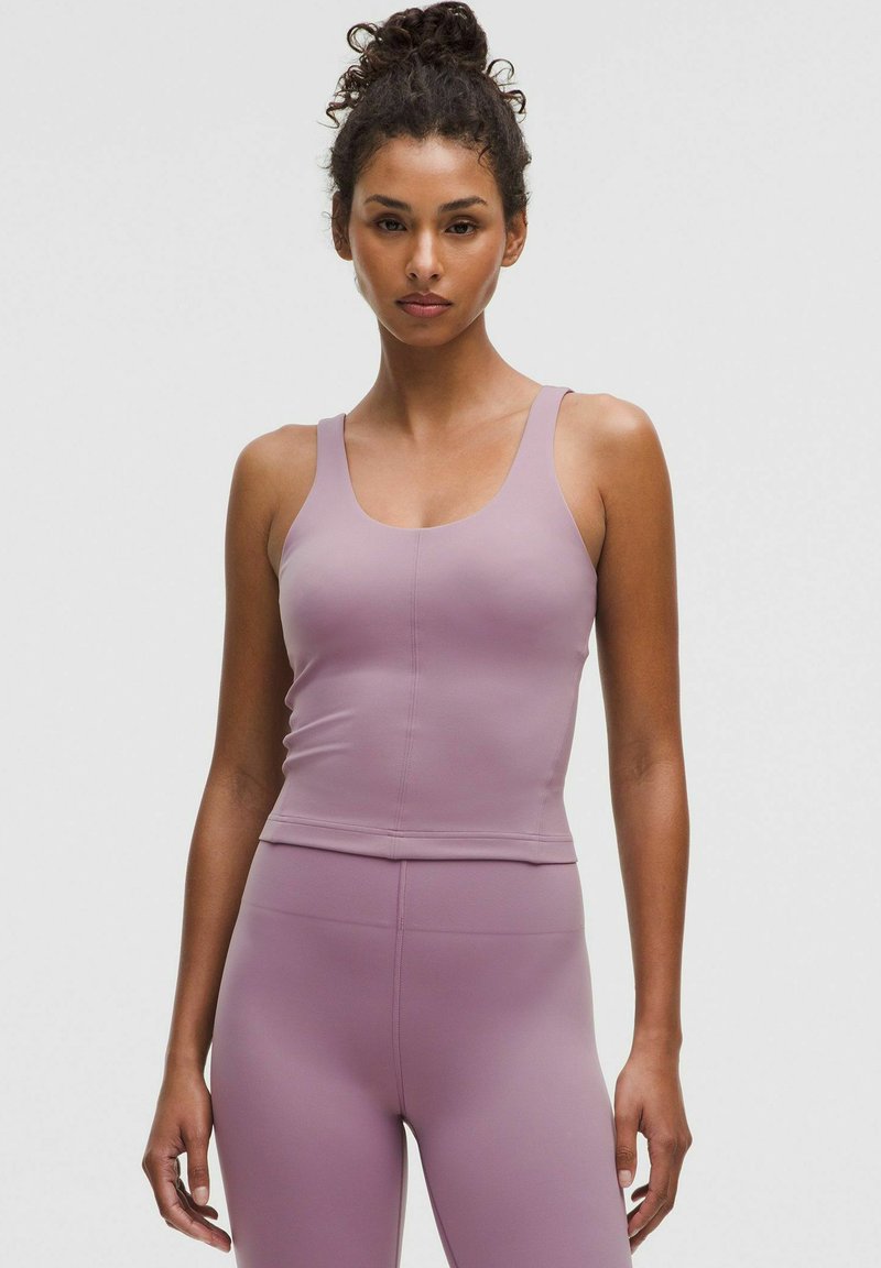 Woman with curly hair in a bun wearing a sleeveless mauve workout top and matching high-waisted leggings standing against a plain background.