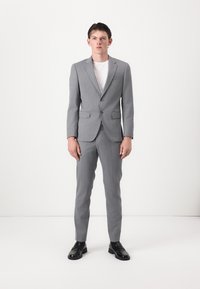 Costume gris sur mesure avec revers cranté, fermeture �à un bouton et deux poches avant. Porté sur un t-shirt blanc et associé à des chaussures noires.