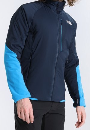 Marineblauwe en blauwe outdoorjas met lange mouwen, ritssluiting aan de voorkant, gestructureerde stof en contrasterende blauwe accenten op de armen en kraag. Opvallend logo op de borst.