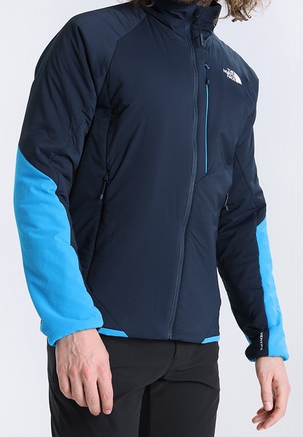 Marinblå och blå utomhusjacka med långa ärmar, dragkedja fram, strukturerad tyg och kontrasterande blå detaljer på armarna och kragen. Anmärkningsbar logotyp på bröstet.