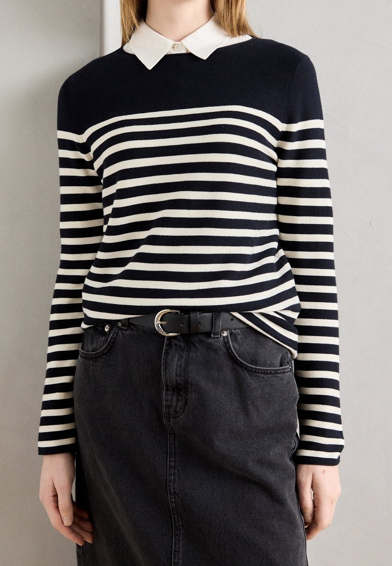 Osoba ubrana w czarno-białą w paski bluzkę z długim rękawem, z białym kołnierzykiem, wsunięta w czarną dżinsową spódnicę oraz czarny pasek z srebrną klamrą.