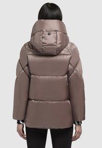 Puffer-takki pehmeässä ruskeassa kankaassa, jossa on suuri huppu ja tikattu kuvio. Hiukan lyhennetty leveillä hihoilla ja piilotetut yksityiskohdat.