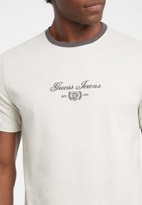 Světle šedé tričko s tmavě šedým ribbed límcem, se vyšívaným logem a emblémem "Guess Jeans" uprostřed.