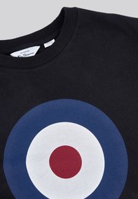 Camiseta de algodón orgánico negra con un gran gráfico circular: anillo exterior azul marino, centro blanco y punto central rojo. Diseño sencillo con cuello redondo.